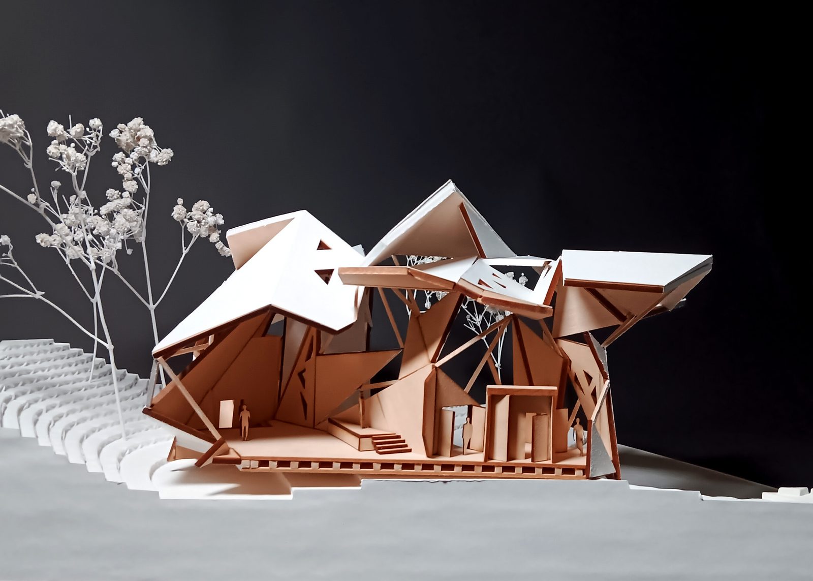 Study model for the Spring 2026 final project: folded white roof planes carried on splayed timber struts over angular chipboard walls, standing on a stepped landscape base with scale figures beneath.